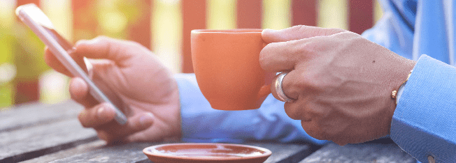 Hands resting on a table holding a mobile phone and a cup of coffee